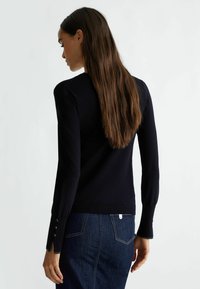 Zwarte gebreide trui met lange mouwen, voorzien van knoopdetails op de manchetten, gecombineerd met donkere spijkerbroek, vanuit een zijaanzicht weergegeven.