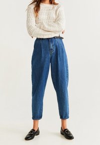 Personne portant un pull blanc en tricot, un jean bleu ample taille haute avec ceinture noire, et des mocassins noirs, debout les bras croisés.