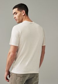 T-shirt corto beige chiaro con maniche corte e colletto a girocollo, realizzato in un tessuto morbido, mostrato da dietro, abbinato a pantaloni verde oliva.