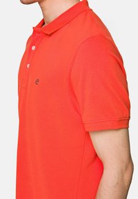 Koszulka polo z krótkim rękawem w jasnopomarańczowym kolorze, wykonana z bawełny, z kołnierzykiem, trzema guzikami oraz małym logo w kolorze granatowym na piersi.