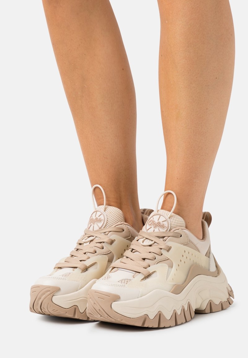 Personne portant des baskets beige et blanc épaisses avec des semelles ondulées, debout sur une surface blanche, montrant des vues de face et de côté.