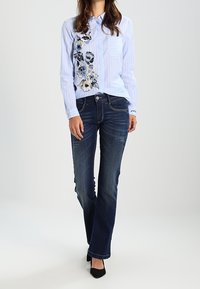 Kobieta w jasnoniebieskiej, pasiasta koszuli z guzikami z haftem kwiatowym, w ciemnoniebieskich dżinsach bootcut, stojąca na białym tle.