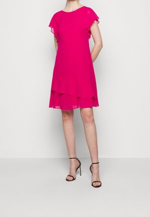 Woman wearing a bright pink, short-sleeve, layered ruffle dress with black ankle-strap high-heeled sandals, standing against a plain background.