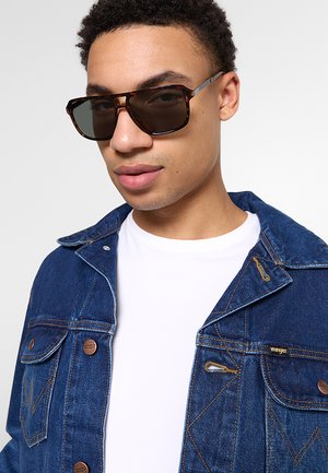 Giovane uomo con occhiali da sole tartarugati scuri, giacca di jeans blu con bottoni in bronzo e maglietta bianca con scollo a girocollo.