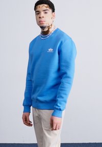 Felpa blu con polsini e orlo a coste, dotata di un piccolo logo bianco sul petto, abbinata a pantaloni chiari. Design semplice.