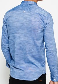 Jasnoniebieska, długozasłonięta koszula z teksturowanej tkaniny. Posiada kołnierzyk zapinany na guziki oraz widoczne detale szwów wzdłuż pleców.
