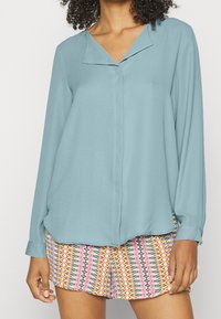 Femme portant un chemisier bleu clair à manches longues avec col et un short à motifs zigzag multicolores.