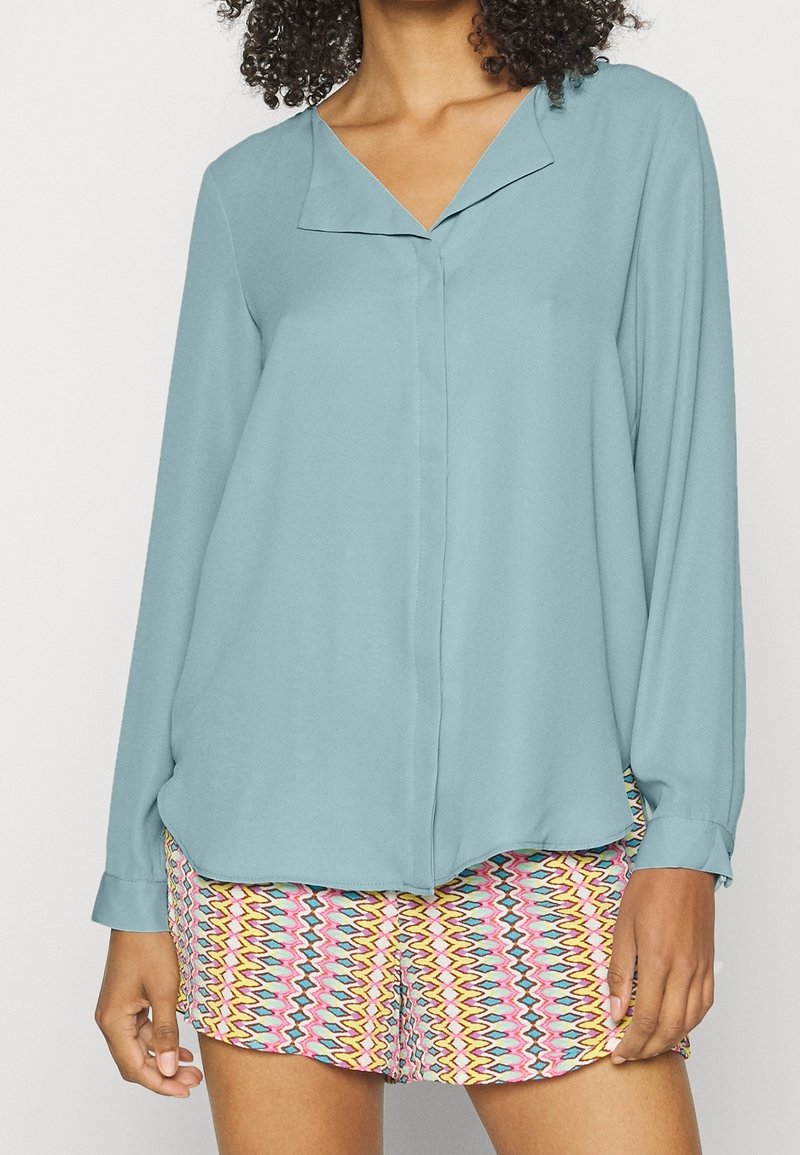 Femme portant un chemisier bleu clair à manches longues avec col et un short à motifs zigzag multicolores.