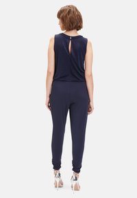 Navyblauer Jumpsuit mit ärmellosem Design, der ein Schlüsselloch-Detail im Rücken und schmal zulaufende Beine hat. Satin-Textur mit glatter Oberfläche.