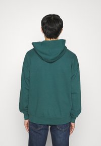 Sudadera con capucha color teal, con un corte holgado, puños y dobladillo de canalé. Textura suave con un acabado liso. Combinada con jeans de color azul oscuro.