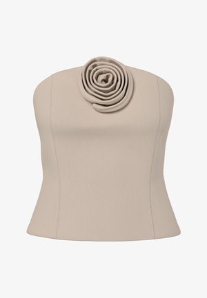 Vrouw met krullend haar, gekleed in een beige strapless top en broekset met rozenvormige details, lopend op een grijze vloer tegen een witte achtergrond.