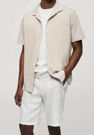 Mężczyzna ubrany w beżową koszulę z krótkim rękawem i guzikami na białą koszulkę oraz białe spodenki do kolan, ręka w kieszeni, neutralne tło.