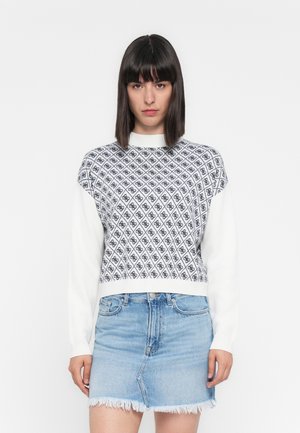 Giovane donna con capelli scuri e lisci, che indossa un maglione bianco con un motivo geometrico nero e una gonna di jeans azzurri chiari con orlo sfrangiato.