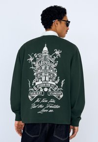 Mørkegrøn sweatshirt med trykt arkitektonisk design og tekst på ryggen. Lange ærmer med ribstrikkede manchetter.