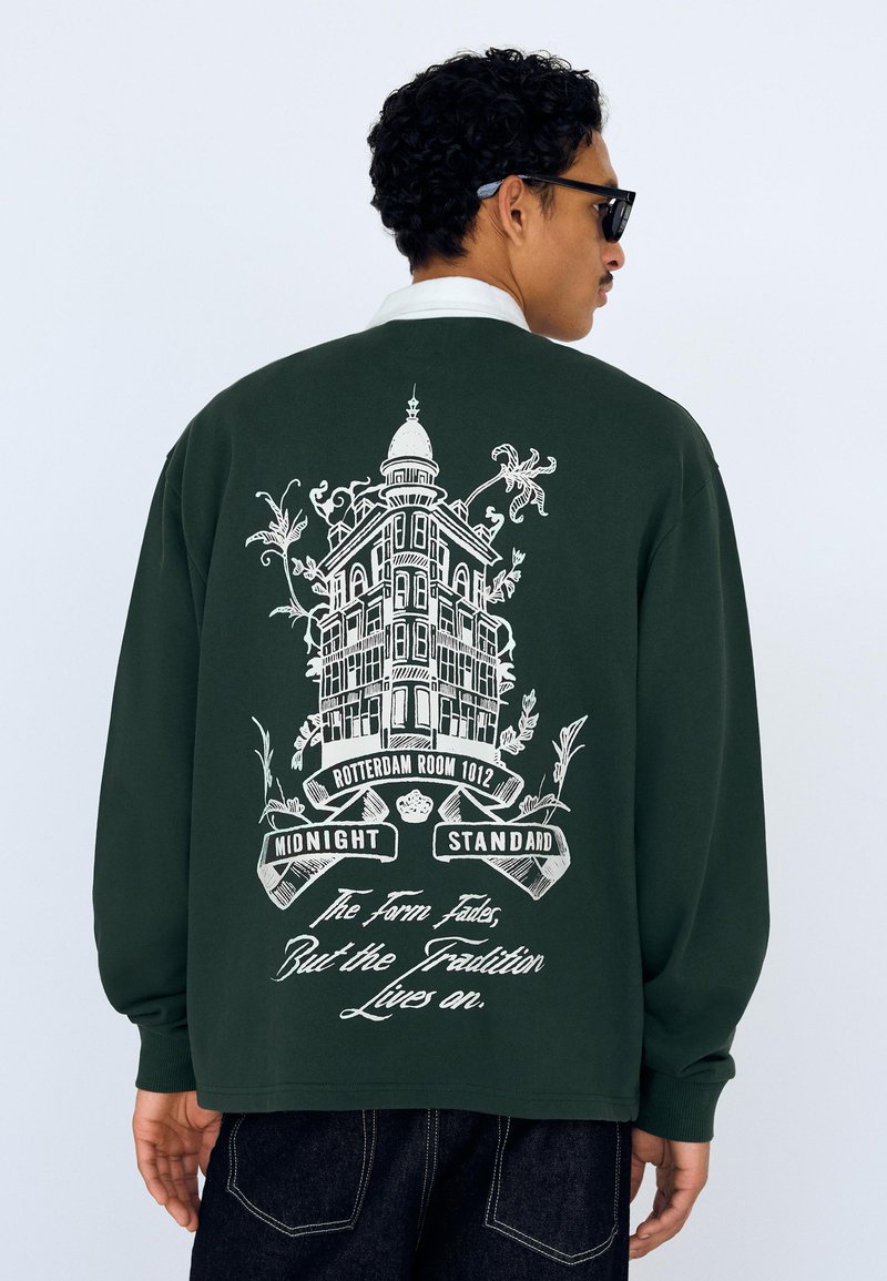 Mørkegrøn sweatshirt med trykt arkitektonisk design og tekst på ryggen. Lange ærmer med ribstrikkede manchetter.