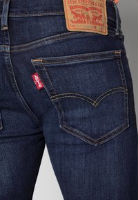 Mörkblå denimjeans med hög midja, guldtråd och ett lädermärke med varumärkeslogotyp på baksidan. Två bakfickor synliga.