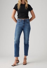 Femme portant un jean bleu taille mi-haute, un t-shirt noir court et des sandales à talons noires à lanières, debout devant un fond uni.
