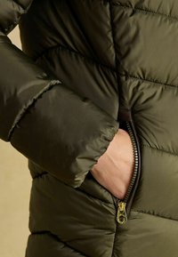 Tmavozelená páperová bunda s prešívanou textúrou, so zipsovým vreckom a lesklým povrchom. Obsahuje priliehajúci golier a výrazné prešívanie.