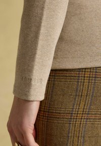 Beige gerippter Langarmshirt mit "Joules"-Stickerei am Handgelenk, kombiniert mit braunen karierten Hosen und einer Hand, die einen goldenen Ring trägt.