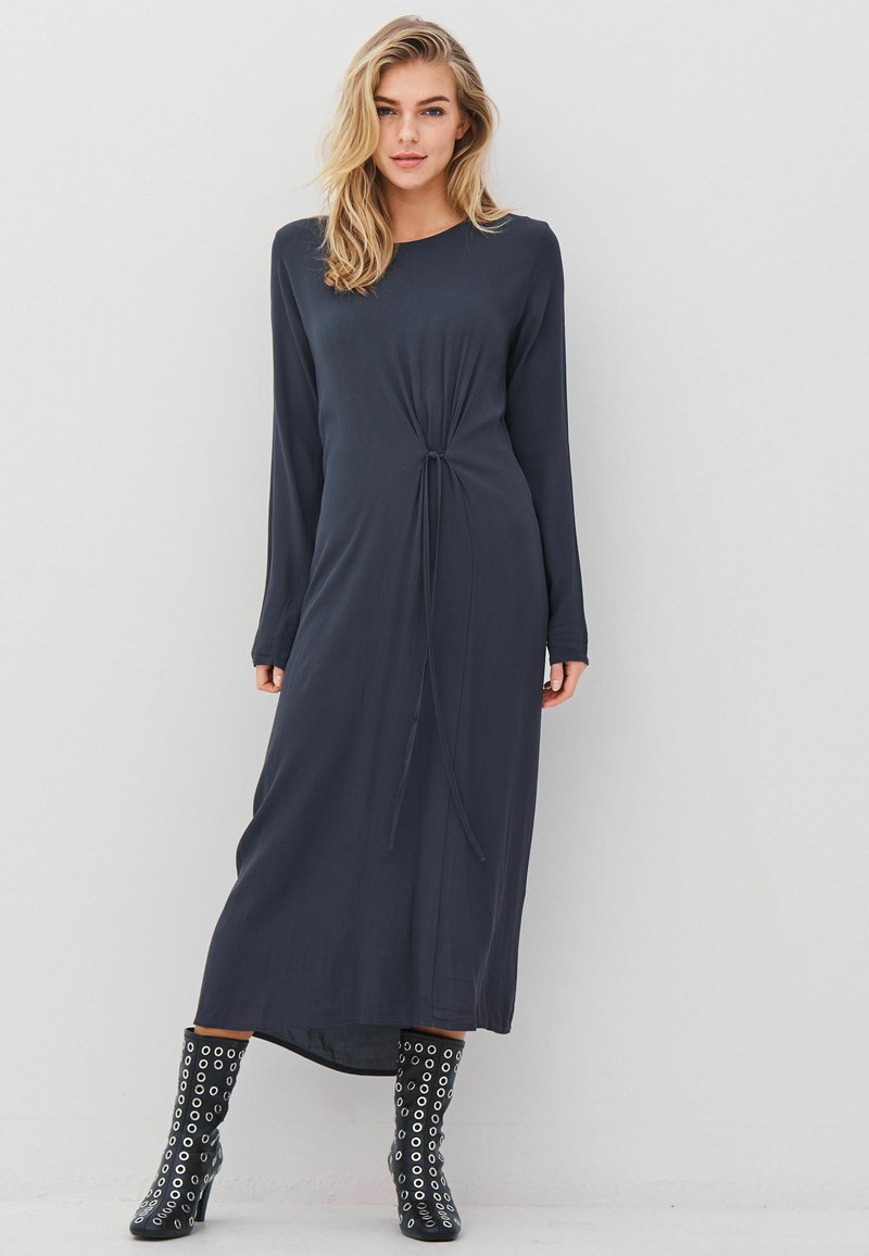 Donna bionda che indossa un abito midi a maniche lunghe di colore blu scuro con dettaglio annodato sul davanti e stivali neri con tacchi alti e occhielli d'argento.