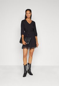 Robe noire à manches trois quarts, taille ceinturée, ourlet en dentelle, associée à des bottes de cow-boy noires avec broderies blanches et un sac à main noir.