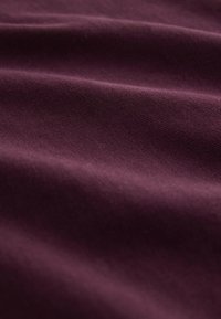 Bordeauxrode stof met een zachte textuur en een lichte glans, die gladde, golvende plooien en een fijn gebreid patroon toont.