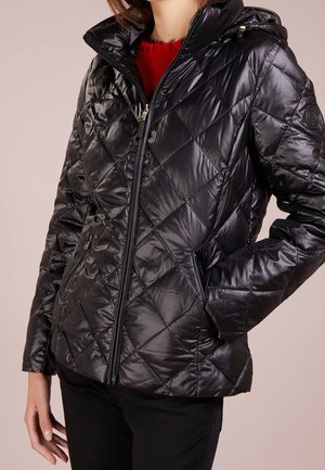 Vrouw draagt een zwarte doorgestikte jas met rits en capuchon over een rode trui, met één hand in de jaszak.