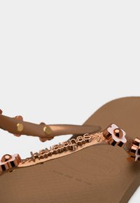 Καφέ σαγιονάρες Havaianas με μοτίφα λουλουδιού σε ροζ χρυσό και ανάγλυφο λογότυπο Havaianas στο λουράκι.