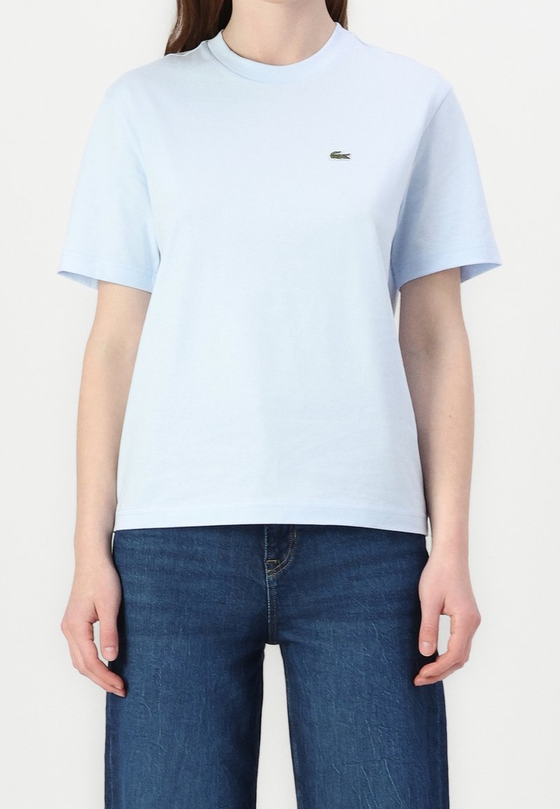 Femme portant un t-shirt à manches courtes bleu clair avec un petit logo de crocodile vert sur la poitrine et un jean bleu foncé.