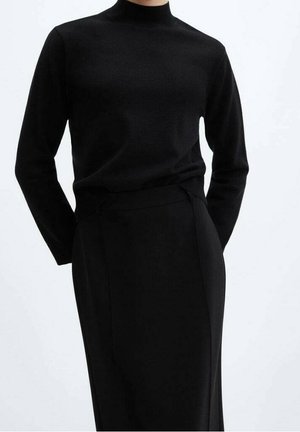 Femme portant un pull noir moulant à col roulé, rentré dans une jupe noire taille haute, sur un fond uni.