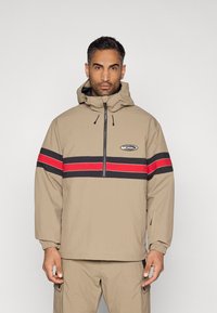 Μπεζ αδιάβροχο με φερμουάρ μπροστά, μαύρες και κόκκινες οριζόντιες ρίγες και ρυθμιζόμενη κουκούλα. Διαθέτει ελαστικούς καρπούς και πλαϊνές τσέπες.