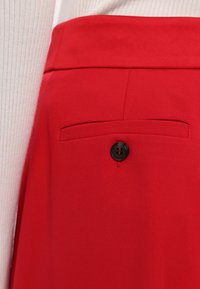 Červené kalhoty na míru s hladkou strukturou, vybavené černým knoflíkem a malou kapsou na zadní straně. Detailní záběr ukazuje detaily.