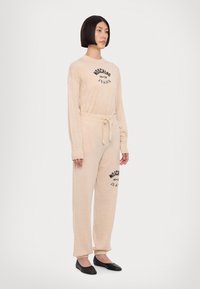 Bežna garnitura za prosti čas z dolgimi rokavi in pasom na vrvico, z črnim potiskanim besedilom. Mehka tekstura z oprijetimi manšeti in zapiranjem okrog gležnjev.