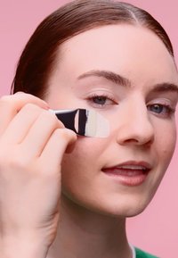 En modell påfører et kremprodukt på kinnet med en flat pensel. Penselen har svart håndtak og hvite børster. Myk rosa bakgrunn.