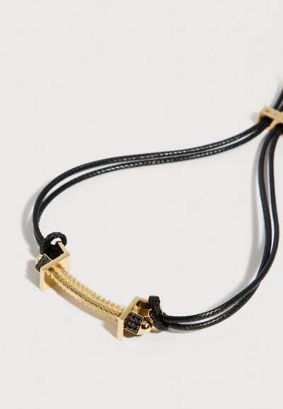 Καμπυλωτό ράβδος σε χρυσό τόνο με μαύρες πέτρες συνδεδεμένη με διπλά μαύρα κορδόνια σχηματίζοντας βραχιόλι σε λευκό φόντο.