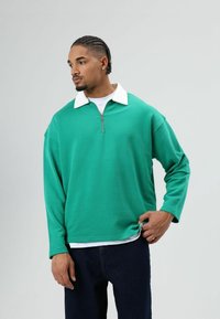 Groene quarter-zip trui met een contrasterende witte kraag, gemaakt van een glad stof. Heeft lange mouwen en een losse pasvorm, gecombineerd met donkere spijkerbroek.