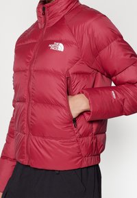 Persona que lleva una chaqueta acolchada roja con el logo de The North Face, con la mano en el bolsillo lateral con cremallera, combinada con pantalones negros.
