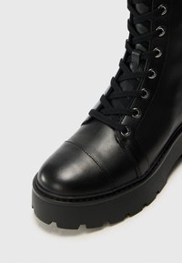 Bottine en cuir noir avec un bout arrondi, à lacets à l'avant, œillets en métal argenté et semelle en caoutchouc noir épaisse. Texture lisse avec des détails de couture.