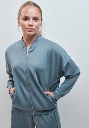 Jeune femme aux cheveux blonds attachés en queue de cheval porte une veste bleu clair zippée et un pantalon assorti, les mains dans les poches.