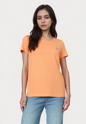 Jeune femme portant un t-shirt orange uni à manches courtes et un jean bleu clair, debout devant un fond blanc.