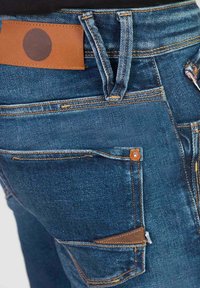 Denimspijkerbroeken in een diepblauwe kleur, met een leren patch op de tailleband en koperen stiksels. Inclusief meerdere zakken en een versterkt ontwerp.