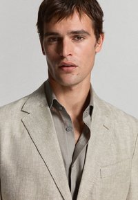 Jeune homme aux cheveux courts et foncés, portant un blazer beige en chevrons par-dessus une chemise grise boutonnée, sur un fond clair uni.