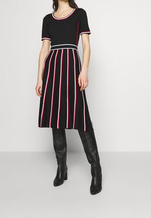 Femme portant une robe noire avec des rayures verticales rouges et blanches et une bordure assortie, associée à des bottes noires hautes à talons, debout contre un mur blanc.