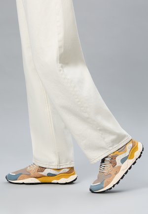 Persona che indossa pantaloni panna e sneakers multicolore con accenti beige, blu, bianchi e gialli che cammina su sfondo grigio.