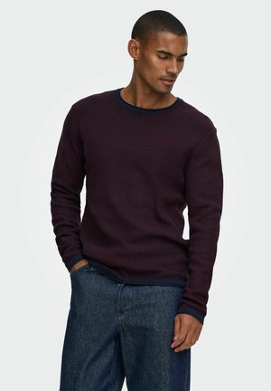 Homme portant un pull violet foncé à manches longues et un jean bleu, une main dans la poche, regardant vers le bas sur le côté devant un fond clair uni.