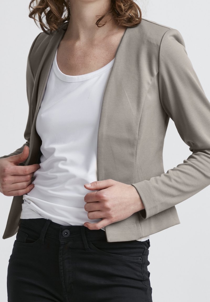 Ljusgrå kavaj med skräddarsydd passform, långa ärmar och öppen front. Bärs över en vit skjorta, ihop med svarta byxor. Slät tygtextur.