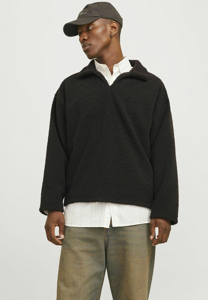Schwarzer Fleece-Pullover mit V-förmigem Kragen, langen Ärmeln und weicher Textur, über einem weißen Hemd mit Knöpfen und einer Jeans getragen.