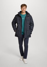 Marinblå vadderad jacka med huva, med flera framfickor, kraftig textur och avslappnad passform. Bärs över en teal-färgad tröja och mörka byxor.