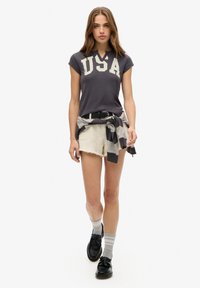Mørkegrå polo shirt med "USA" i hvid, parret med beige denim shorts. Accentueret med en stribet trøje bundet om taljen. Sorte sko og grå sokker.
