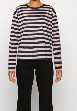 Personne portant un pull à manches longues à rayures horizontales bleu marine et rose clair avec un pantalon noir, se tenant devant un fond clair uni.